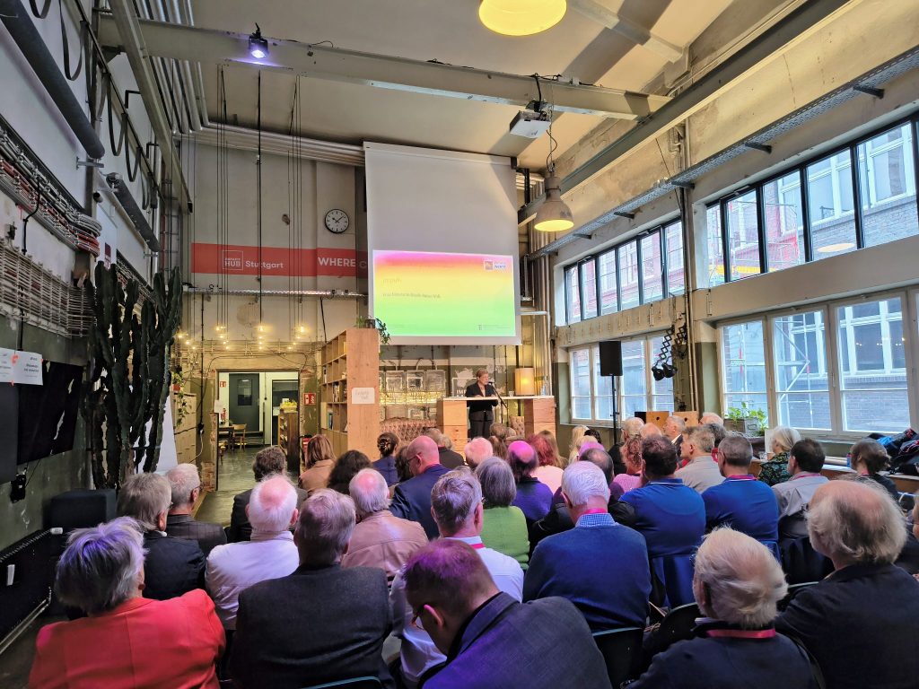 Das Foto zeigt eine dynamische Barcamp-Session im hellen Konferenzraum mit 20-30 Teilnehmern in lockeren Gruppen um runde Tische. Im Vordergrund Dr. Diana Wiedemann von Grundmann + Wiedemann diskutiert lebhaft mit Ehrenamtlichen; Flipcharts mit Stichworten „Vernetzung", „Weiterbildung", „DenkmalnetzBW" sind sichtbar. Hinten Ministerin Nicole Razavi am Podium, dahinter Banner des Landesamts für Denkmalpflege BW und Stiftung BauKulturerbe, natürliches Tageslicht durch große Fenster.