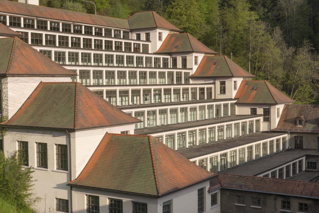 Junghans Terrassenbau Schramberg Industriedenkmal – Museum Schwarzwald Uhren Philipp Jakob Manz. - Historisches Gebäude mit Terrassenstruktur - Das Bild zeigt den ikonischen Junghans-Terrassenbau in Schramberg, ein denkmalgeschütztes Industriedenkmal aus 1916–1918 vom Architekten Philipp Jakob Manz. Die terrassierte Hangbebauung mit neun Etagen, durchgehenden Fensterbändern und Treppenhäusern diente ehemals der Uhrenproduktion und beherbergt heute das Junghans Terrassenbau Museum.